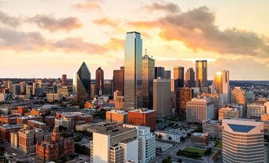 Aerial view of downtown Dallas, Texas at sunset, featuring a mix of modern skyscrapers and historic buildings under a partly cloudy sky with warm golden light.