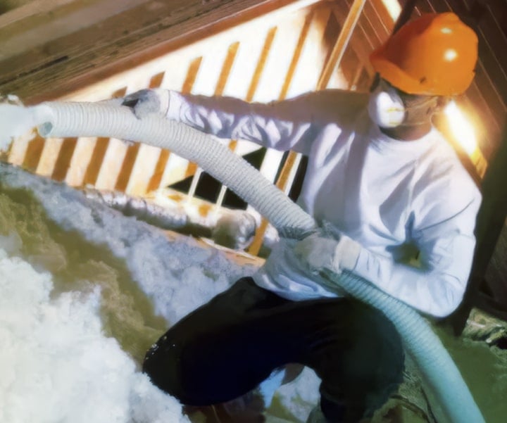 A worker in a hard hat, face mask, and gloves is using a large hose to spray loose-fill insulation into an attic space. The surrounding area is already covered with thick white insulation, and the attic framing is visible in the background.