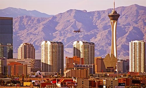 Las Vegas city skyline at sunset with the Stratosphere Tower prominently in view, surrounded by tall hotels and residential buildings, with rugged desert mountains in the background and a helicopter flying above the city.