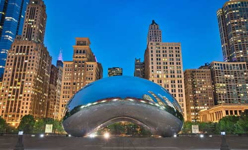 The Cloud Gate sculpture, commonly known as "The Bean," reflects the surrounding city skyline at dusk in Millennium Park, Chicago. Tall buildings frame the metallic sculpture, which mirrors the lights and architecture around it under a deep blue sky.