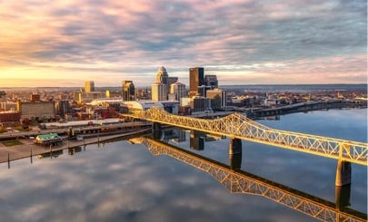 A panoramic view of downtown Louisville, Kentucky at sunrise, featuring the skyline reflected in the Ohio River. The prominent bridge spans the calm water, connecting the city and adding symmetry to the serene scene.