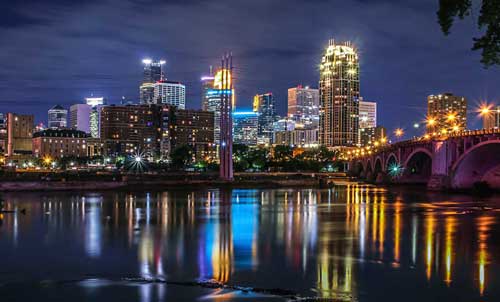Downtown Minneapolis skyline at night with illuminated buildings reflecting vibrant colors on the Mississippi River, alongside a lit-up bridge to the right.