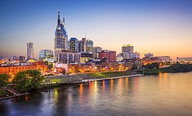 Nashville skyline at dusk featuring the AT&T Building, known as the “Batman Building,” with city lights reflecting on the Cumberland River and a colorful evening sky in the background.