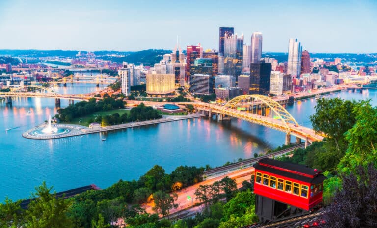 Scenic view of downtown Pittsburgh at sunset, showing the city skyline, yellow bridges spanning the rivers, the Point State Park fountain at the river confluence, and the iconic red Duquesne Incline cable car ascending the green hillside in the foreground.