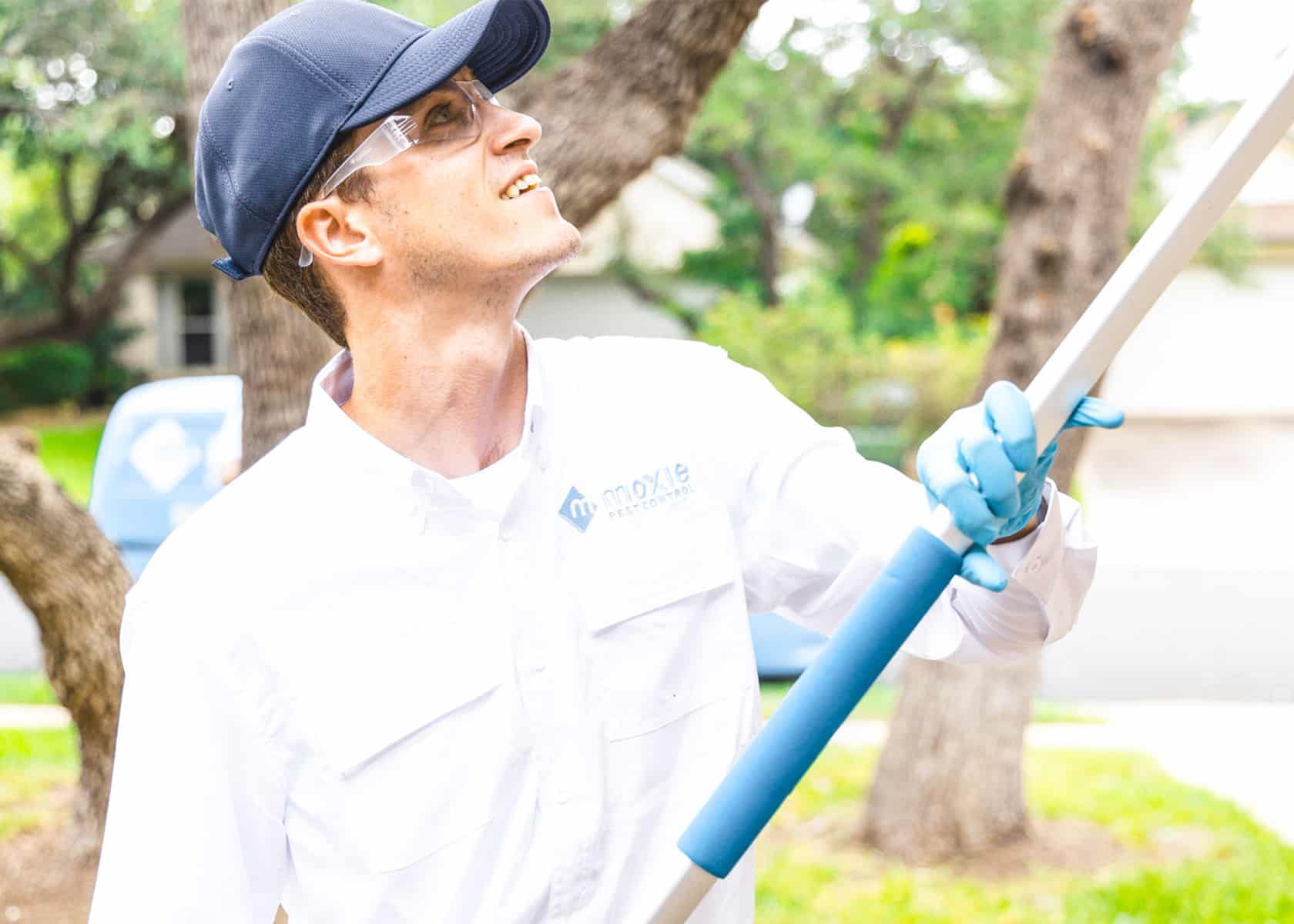 A pest control technician in a white uniform shirt with a "Moxie Pest Control" logo uses a long tool while looking upward. He wears a navy cap, safety glasses, and blue gloves, standing outdoors near trees and a blue service vehicle.