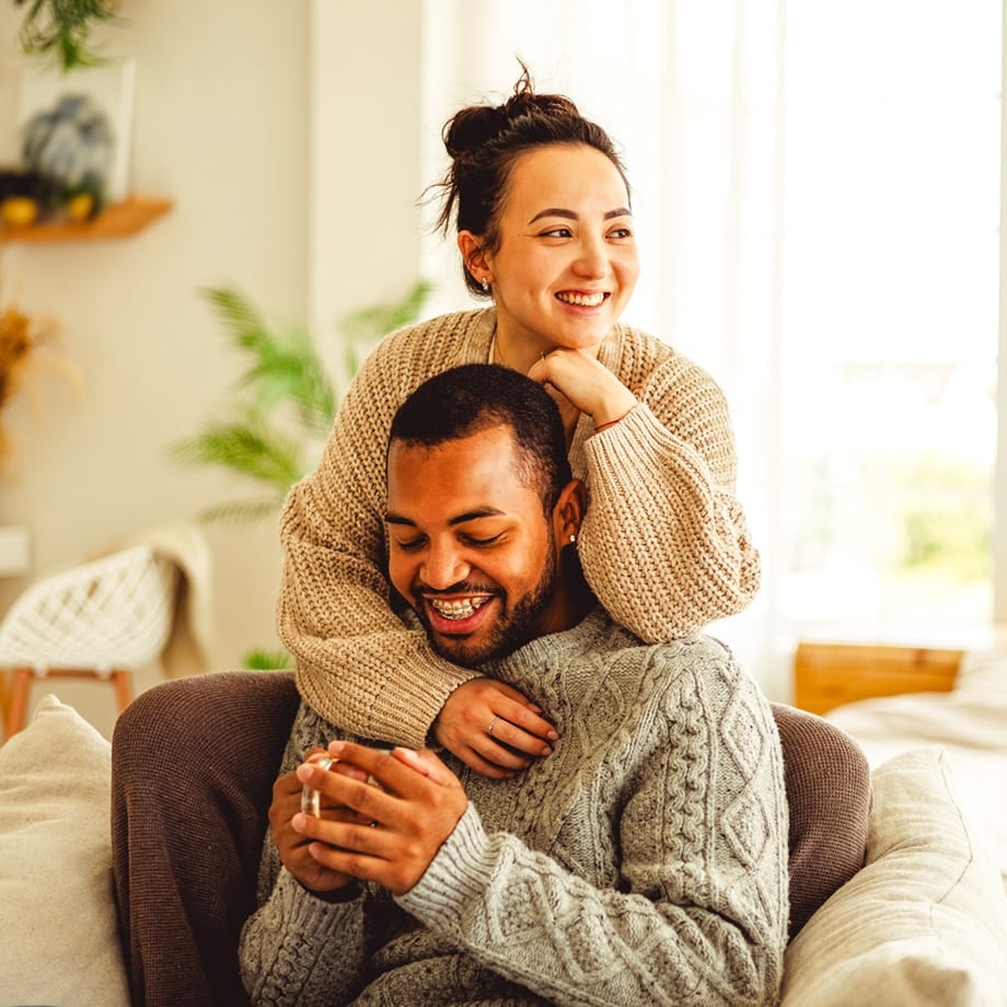 A smiling couple relaxes together in a bright, cozy living room. The man, seated in a chair wearing a gray cable-knit sweater, holds a cup while the woman hugs him from behind, resting her chin on his head and wearing a beige sweater.