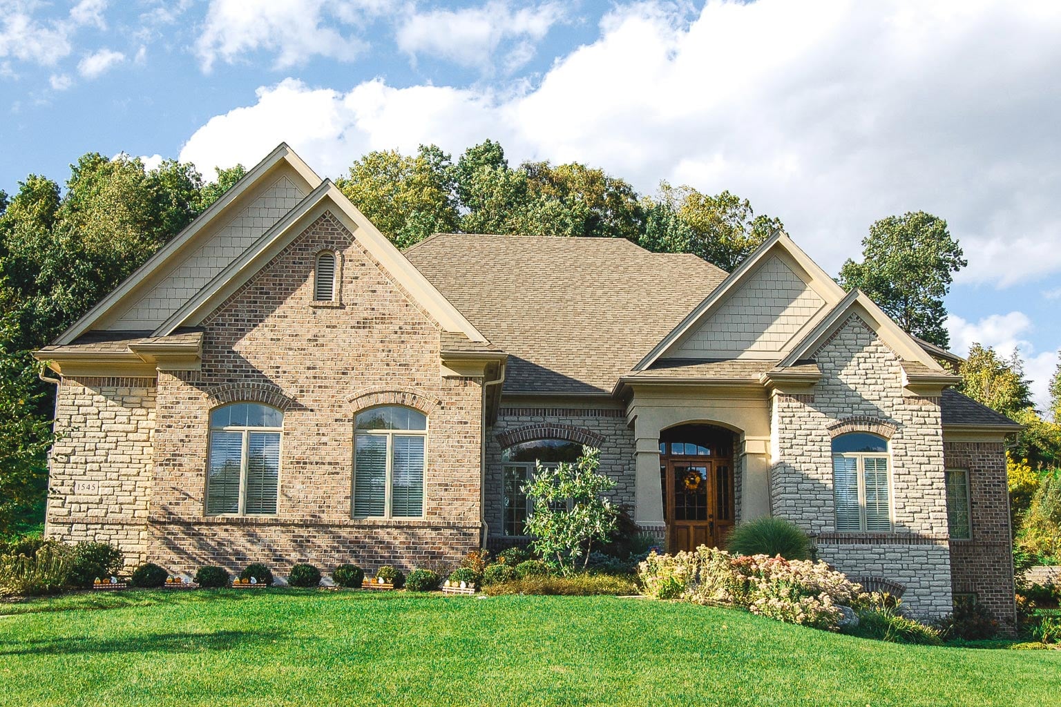 A spacious single-story house with a combination of stone and brick exterior, gabled roof, and large arched windows sits on a neatly manicured lawn. Shrubs and flowering plants line the walkway leading to a wooden front door, framed by a backdrop of tall trees and a partly cloudy sky.