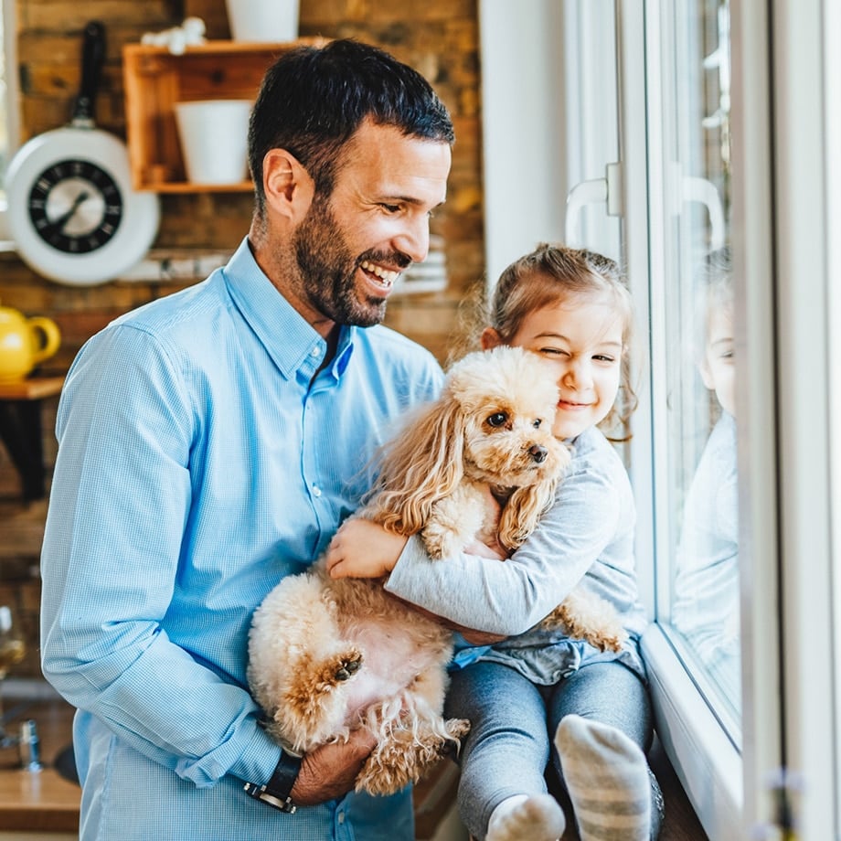 A smiling man in a light blue shirt holds a young girl and a fluffy light-brown poodle as they look out a bright window. The girl, dressed in grey, grins while hugging the dog, and the cozy kitchen setting behind them adds to the homey feel.