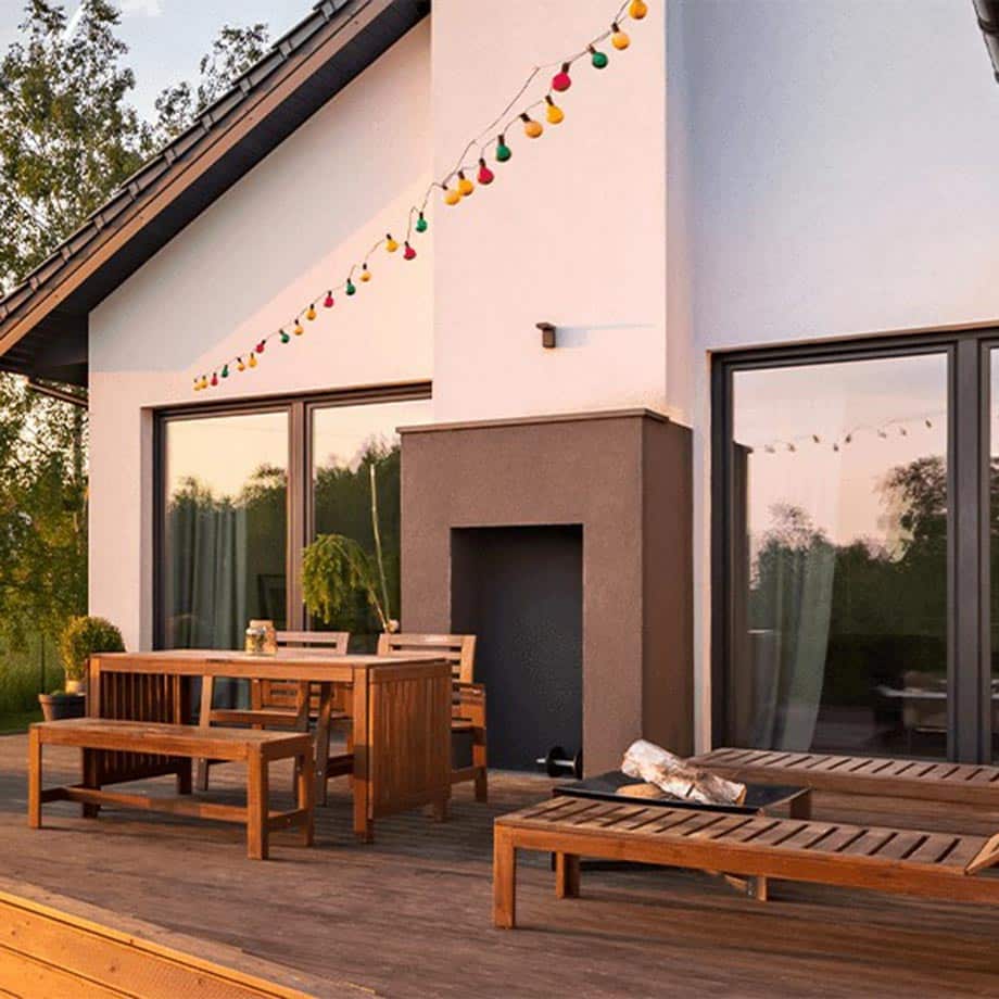 A stylish backyard patio with wooden furniture, including a dining table set and two lounge chairs, sits in front of a modern white house with large glass doors. String lights with colorful bulbs hang above, and a built-in outdoor fireplace adds a cozy touch.